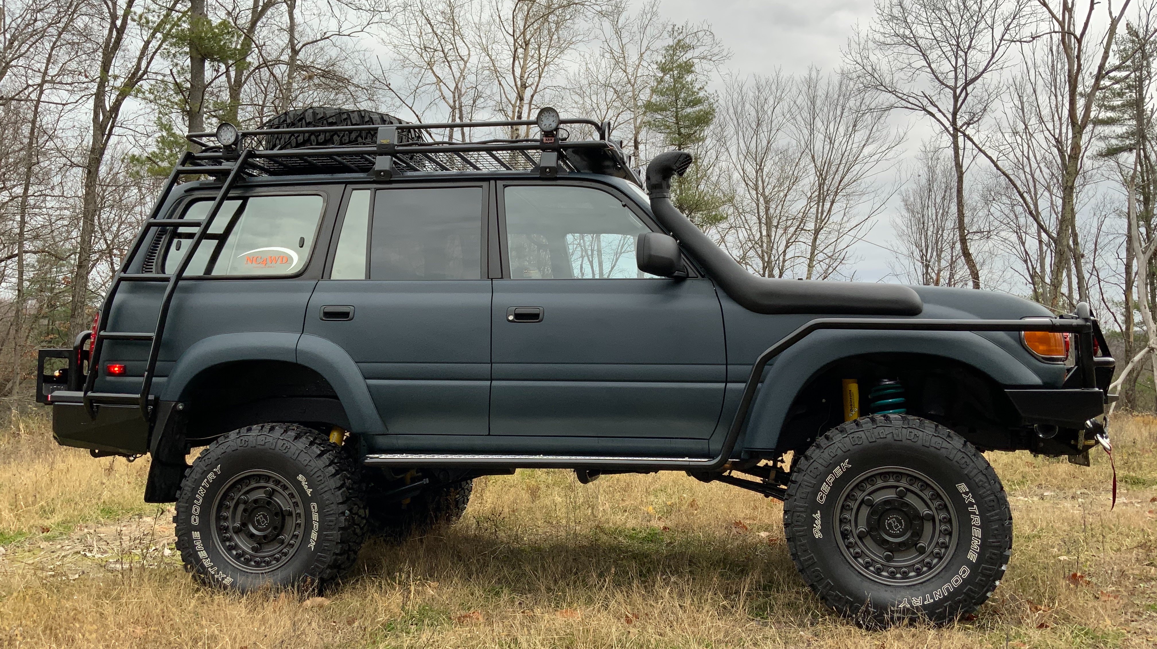 North Country 4WD's dark off-road SUV with a roof rack, snorkel, and oversized tires, parked beside the woods